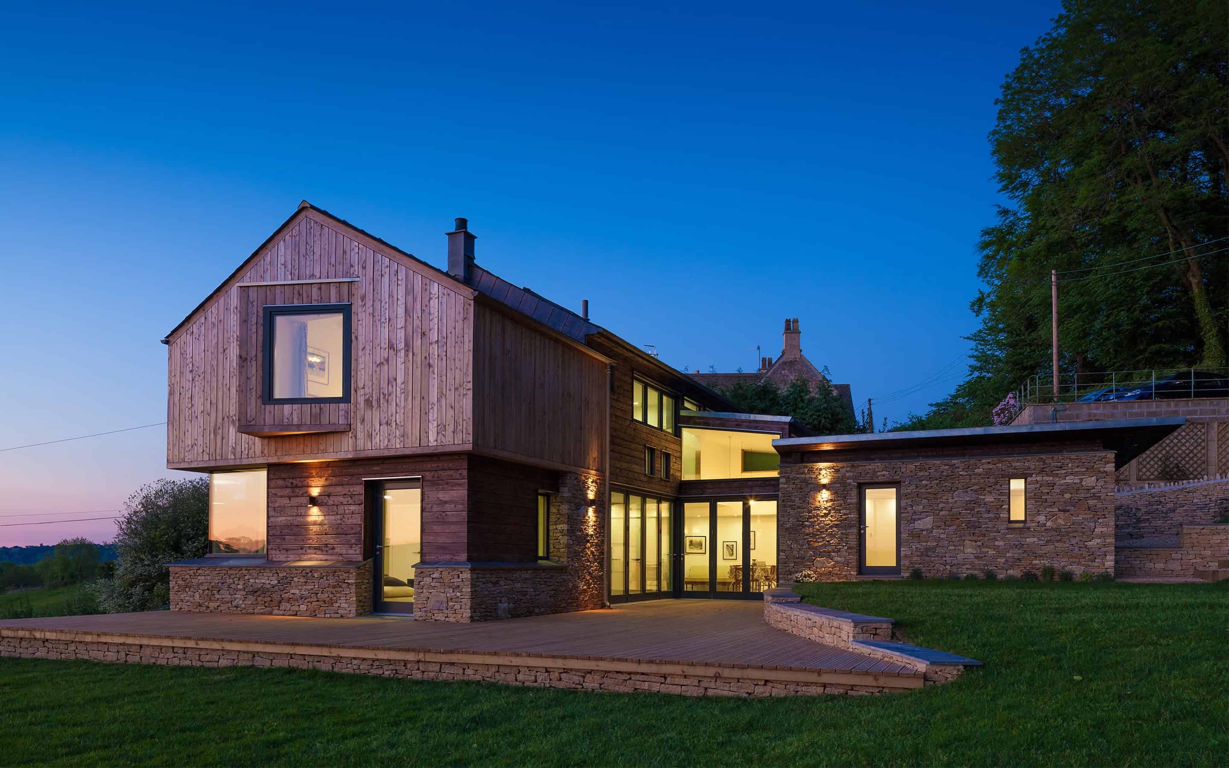 The Larch House full exterior at twilight with larch and stone facade and illuminated living spaces